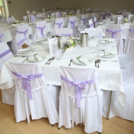 Ein Bankettsaal ist mit mehreren Tischen eingerichtet, die mit weißer Tischdecke und lila Bändern verziert sind. Tischgedecke umfassen Blumen und silberne Karaffen. Der Raum hat Holzböden und eine neutrale Farbgebung.