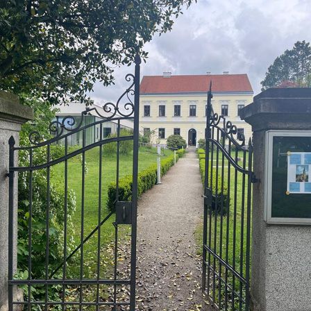 Ein breiter Kiesweg führt zu einem kunstvollen Tor vor einem großen Gebäude mit rotem Dach. Das Tor wird von einem Steinpfeiler mit einem Schild flankiert. Hinter dem Tor sind gepflegte Hecken und grüner Rasen zu sehen.