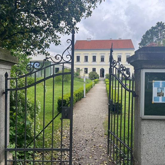 Ein breiter Kiesweg führt zu einem kunstvollen Tor vor einem großen Gebäude mit rotem Dach. Das Tor wird von einem Steinpfeiler mit einem Schild flankiert. Hinter dem Tor sind gepflegte Hecken und grüner Rasen zu sehen.