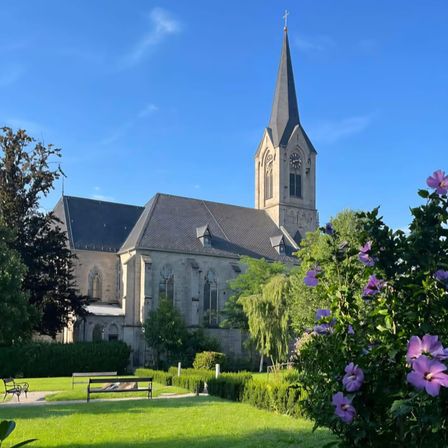 Eine Kirche mit einem spitzen Turm und einer Uhr steht an einem sonnigen Tag. Sie hat mehrere Bogenfenster und ein Kreuz auf der Spitze. Sie ist von einem Rasen und Büschen mit lila Blumen umgeben.
