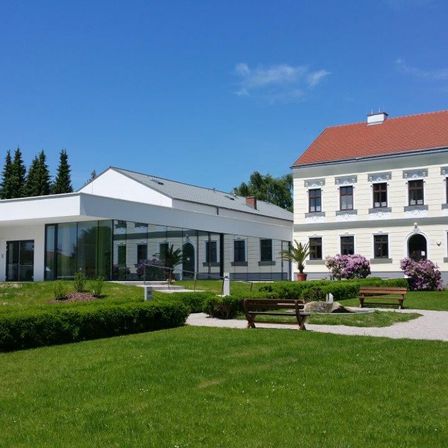 Ein modernes Gebäude mit rotem Dach und mehreren Fenstern steht neben einem älteren Gebäude. Ein grüner Rasen mit zwei Holzbänken und einigen Pflanzen und Bäumen davor.