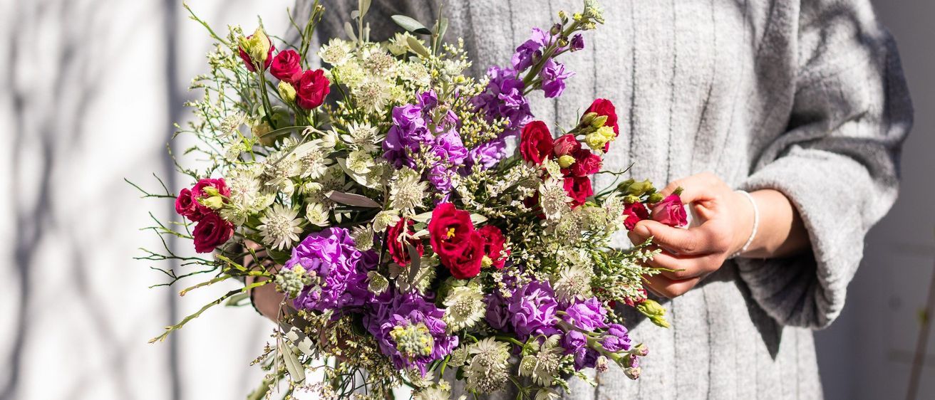 Eine Frau hält einen Blumenstrauß mit einer Mischung aus lila, roten und weißen Blüten. Der Strauß sieht natürlich aus, mit sichtbaren Blättern und Stielen.