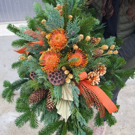 Eine Frau hält eine dekorative Blumenarrangement mit orangen Blumen und Tannenzapfen, vor einem neutralen Hintergrund.