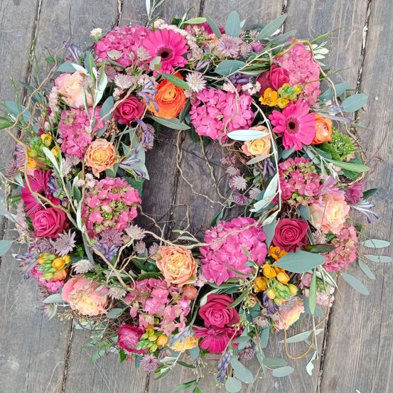 Ein farbenfroher Kranz aus rosa, orange und lila Blumen auf einer Holzoberfläche.