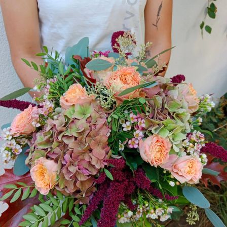 Eine Frau hält einen großen Blumenstrauß mit Rosen, Hortensien und Grünpflanzen. Der Strauß ist farbenfroh mit Rosa-, Lila- und Grüntönen.