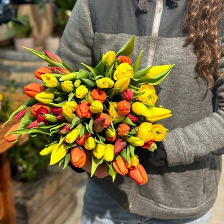 Eine Person in einem grauen Jacke hält einen Strauß gelber und orangefarbener Tulpen. Der Strauß hat grüne Blätter und wird nah am Körper gehalten.