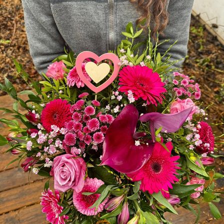Eine Frau hält einen rosa-lila Blumenstrauß mit einem Herz oben drauf. Der Strauß steht auf einer Holzoberfläche.