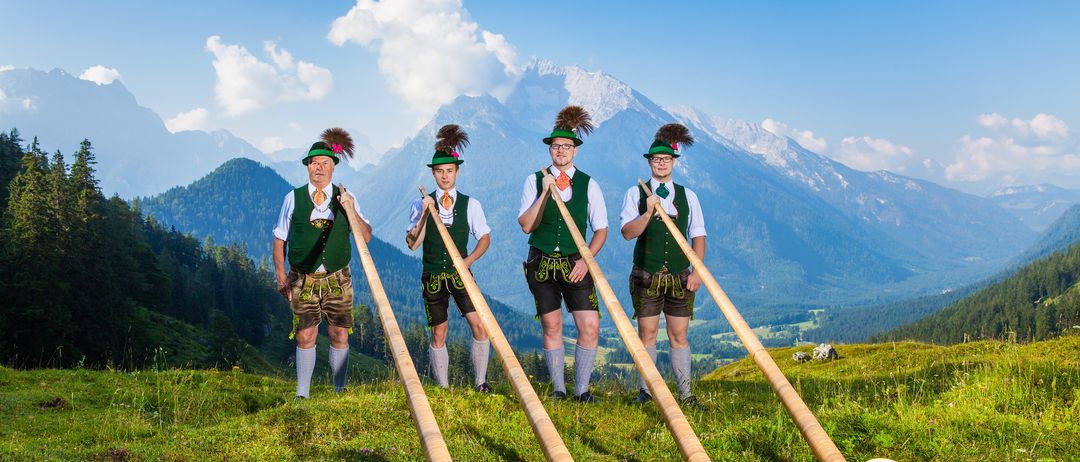 Vier Männer in traditioneller grüner Kleidung und Hüten stehen in einer Reihe, jeder hält eine lange Holztrompete. Sie stehen auf einer grasbewachsenen Anhöhe mit Bergen im Hintergrund.