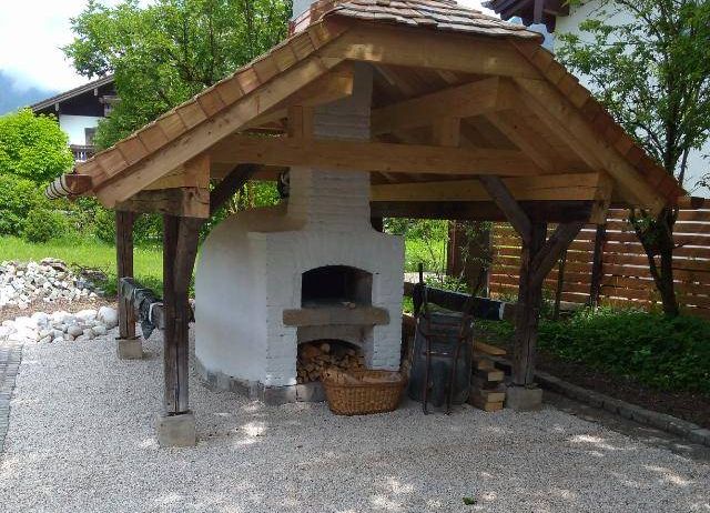 Ein rustikales Holzpavillon mit einem weißen Backofen, einem Kupferdach und einem Schornstein. Es ist von Kies umgeben und in der Nähe befindet sich ein Korb mit Holz.