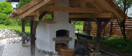 A rustic wooden gazebo with a white brick oven, a copper roof, and a chimney. It is surrounded by gravel and has a basket of wood nearby.