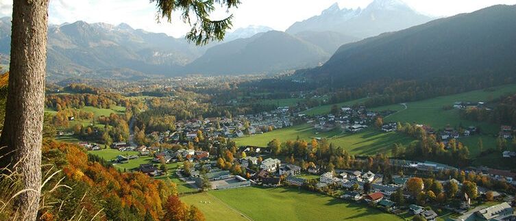 Luftaufnahme eines Dorfes in einem Tal, umgeben von grünen Feldern und bewaldeten Hügeln unter einem blauen Himmel mit Wolken. In der Ferne schneebedeckte Berge.