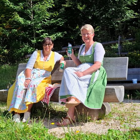 Zwei Frauen in traditioneller Kleidung sitzen auf einer Bank im Park, lächeln und halten Getränke. Hinter ihnen umgibt der Bereich ein Holzzaun und üppige Grünflächen.