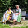Two women in traditional attire sit on a bench in a park, smiling and holding drinks. Behind them, a wooden fence and lush greenery surround the area.