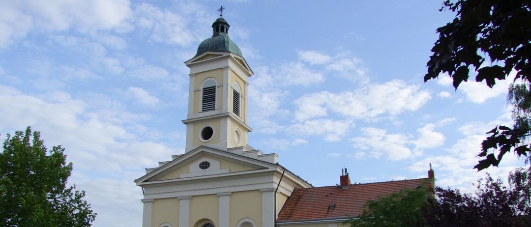 Eine traditionelle gelbe Kirche mit einem grünen Turm und einem roten Dach. Vor dem Gebäude steht ein großer Baum.