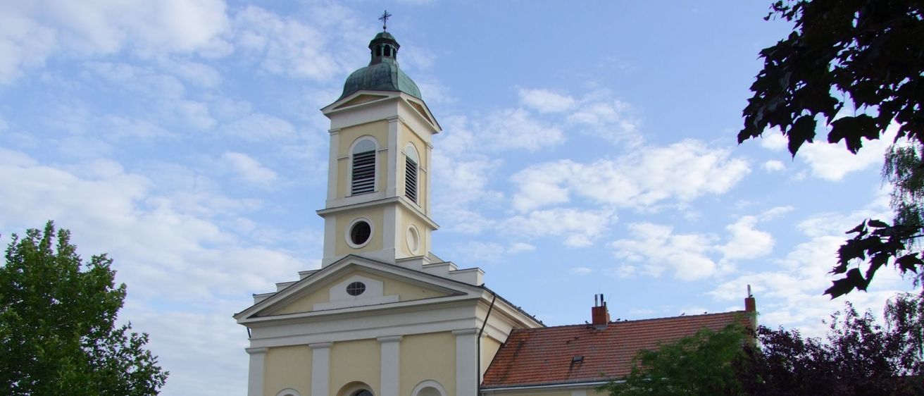 Eine traditionelle gelbe Kirche mit einem grünen Turm und einem roten Dach. Vor dem Gebäude steht ein großer Baum.
