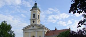 Eine traditionelle gelbe Kirche mit einem grünen Turm und einem roten Dach. Vor dem Gebäude steht ein großer Baum.