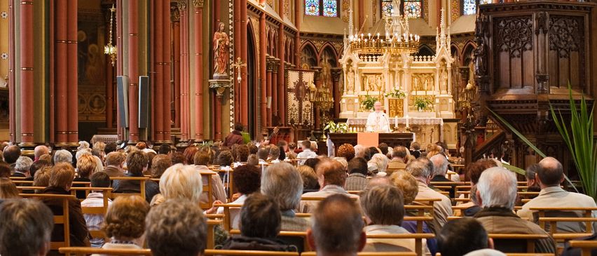 Eine große Gemeinde besucht einen Gottesdienst in einer großartigen Kathedrale, wobei der Priester vor einem goldenen Kronleuchter am Altar steht.