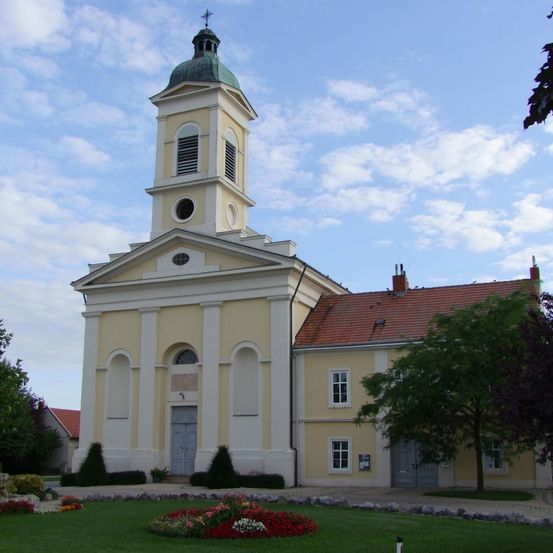 Eine Kirche mit einer grünen Kuppel, einem Glockenturm und einem roten Dach. Das Gebäude ist von einem gepflegten Rasen mit bunten Blumen umgeben.