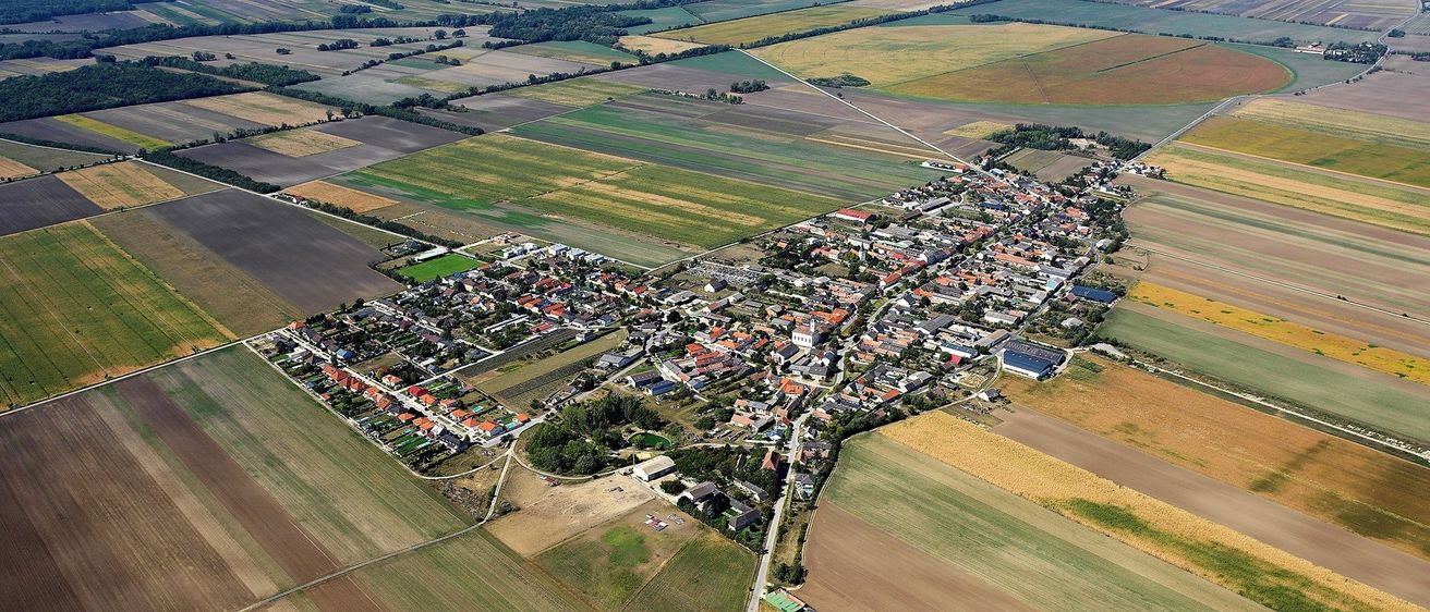 Luftaufnahme einer ländlichen Stadt mit vielen Häusern und Gebäuden, umgeben von großen landwirtschaftlichen Feldern. Grüne und braune Felder sind sichtbar.