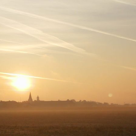 Eine friedliche Landschaft mit einer aufgehenden Sonne über einer Stadt mit einem Turm, die lange Schatten und eine neblige Atmosphäre wirft.
