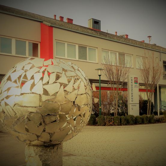 Vor einem Gebäude mit einem roten Streifen, mehreren Fenstern und einem Schild, das Gemeindeamt steht, befindet sich eine große, reflektierende Metallkugel.