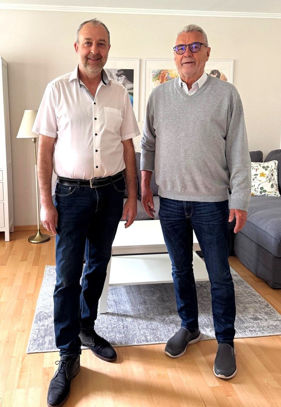 Zwei Männer stehen in einem Wohnzimmer. Einer trägt ein weißes Hemd und Jeans, der andere ein graues Sweatshirt und Jeans. Sie lächeln beide.