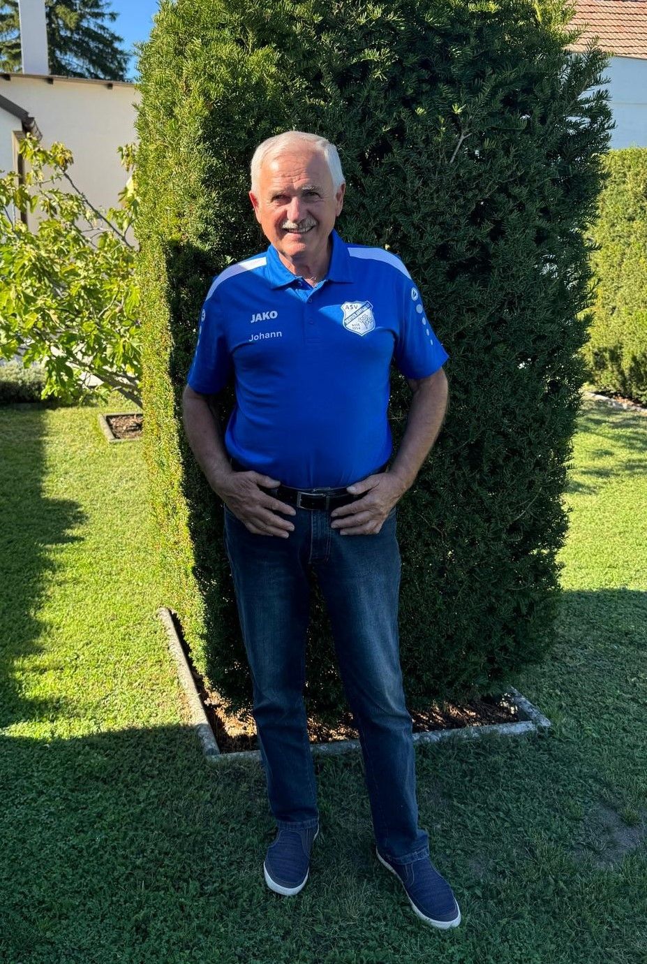 Ein Mann in einem blauen Poloshirt mit dem Namen Johann steht in einem Garten. Er hat ein Lächeln im Gesicht und die Hände in den Hüften.