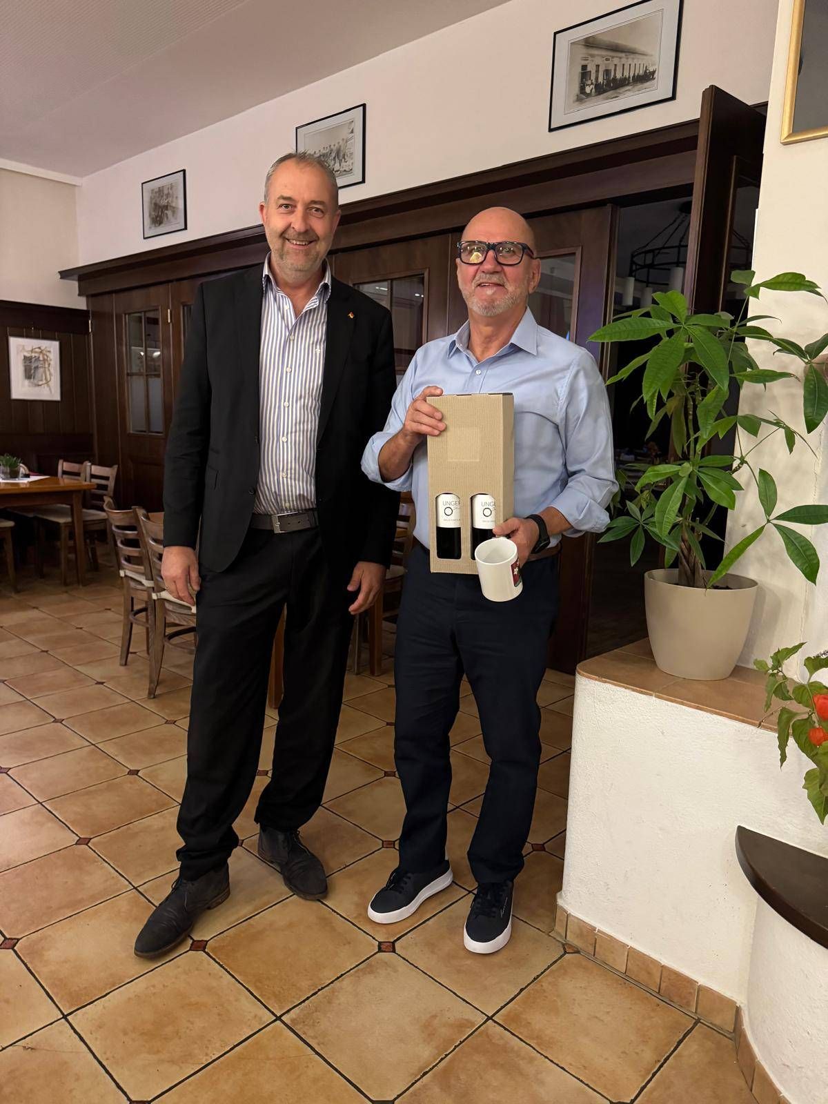 Zwei Männer in formeller Kleidung stehen nebeneinander, einer hält eine Weinschachtel. Sie lächeln und posieren für ein Foto in einem Raum mit Holzboden und einem Topfpflanzen.