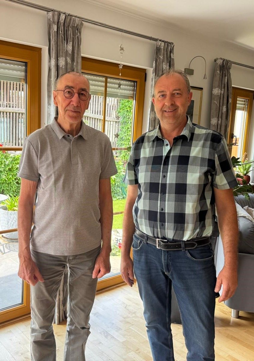 Zwei Männer stehen in einem Raum mit einer Glastür, einer trägt eine Brille und ein graues T-Shirt, der andere ein kariertes Hemd und Jeans. Sie lächeln und schauen in die Kamera.