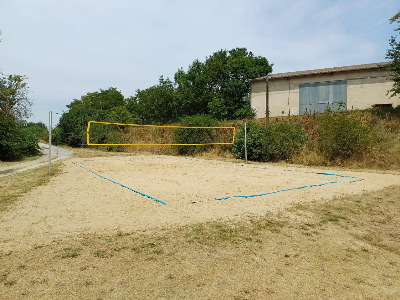 Ein Volleyballfeld im Freien mit einem gelben Netz auf einem sandigen Bereich, umgeben von Grünflächen und einem Gebäude im Hintergrund.