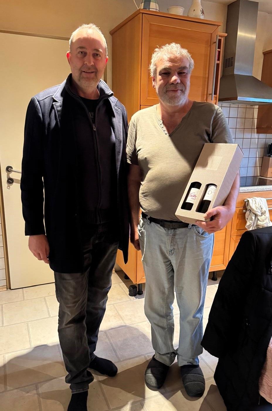 Zwei Männer stehen in einer Küche, einer hält eine Schachtel mit Weinflaschen und lächelt für ein Foto.