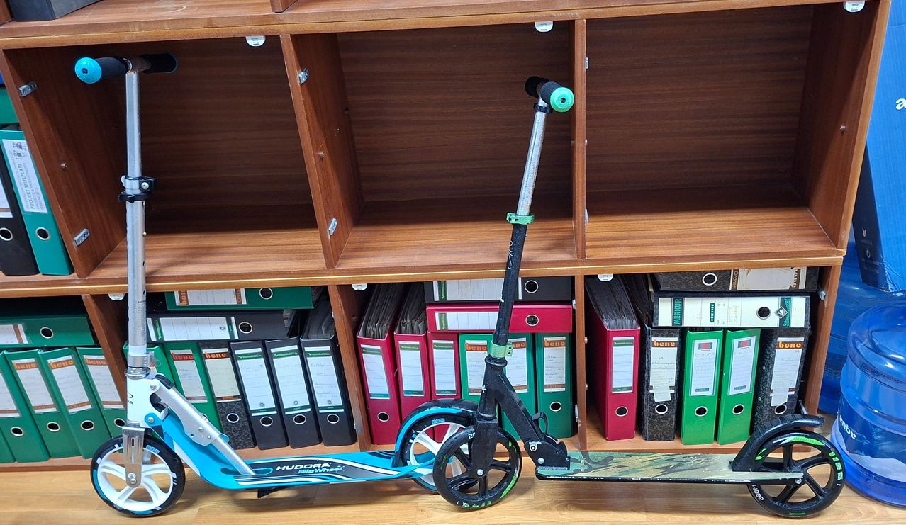 Ein Roller steht vor einem Holzregal mit Fächern und Ordnern und zeigt einen blau-schwarzen Roller mit grünem Griff.