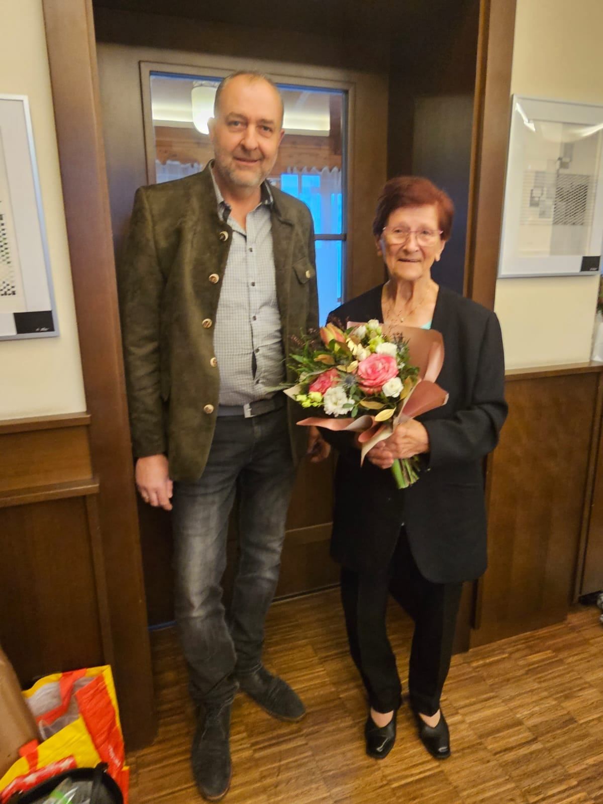 Ein Mann und eine Frau stehen lächelnd vor einer Holztür. Der Mann trägt eine Jacke und Jeans, die Frau einen schwarzen Blazer und eine Brille. Sie hält einen Blumenstrauß. Hinter ihnen befindet sich eine Wand mit gerahmten Bildern.