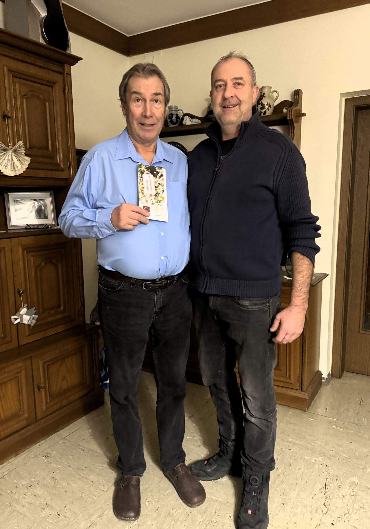 Zwei Männer stehen nebeneinander. Der Mann auf der linken Seite hält eine Karte und lächelt. Hinter ihnen steht ein hölzerner Schrank mit Bilderrahmen und anderen Gegenständen.