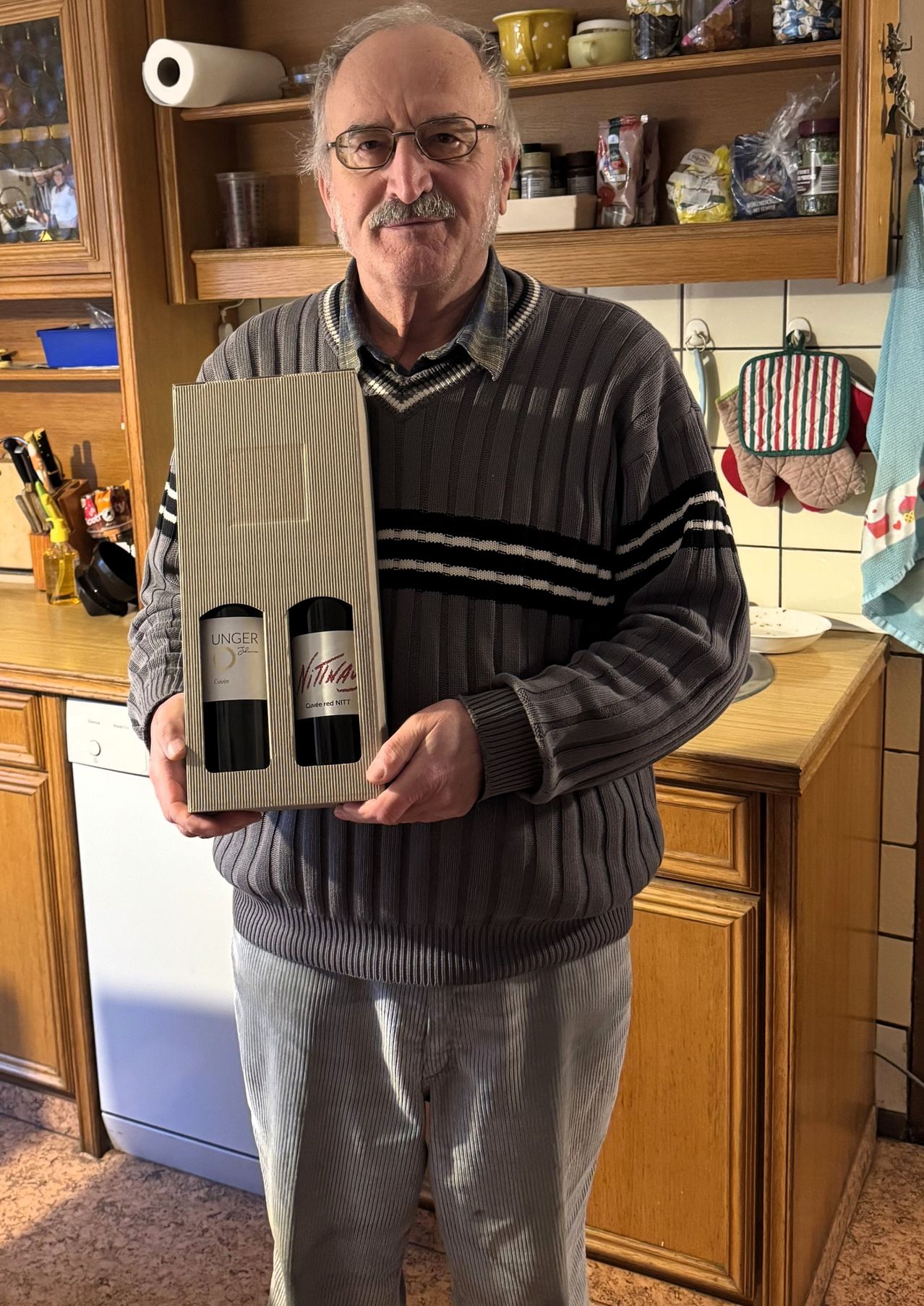 Ein Mann steht in einer Küche und hält eine Schachtel mit zwei Flaschen Unger Wein. Er trägt einen grauen Pullover mit Streifen. Hinter ihm befindet sich eine Holztheke und ein Waschbecken.