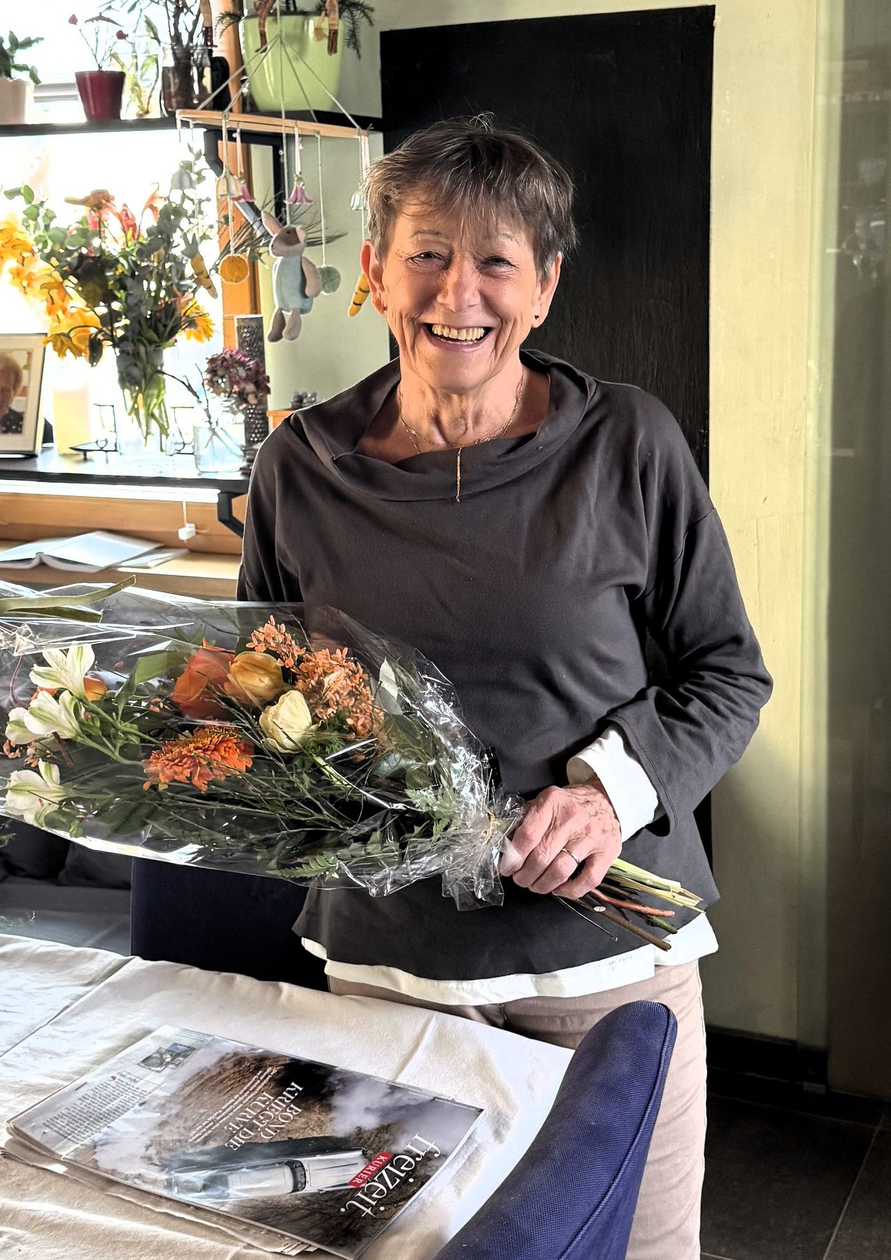 Eine lächelnde Frau hält einen Blumenstrauß. Sie trägt eine graue Kapuzenjacke und eine Halskette. Ein Sofa und ein Tisch stehen hinter ihr. Ein gerahmtes Bild und Blumen sind auf einer Fensterbank.