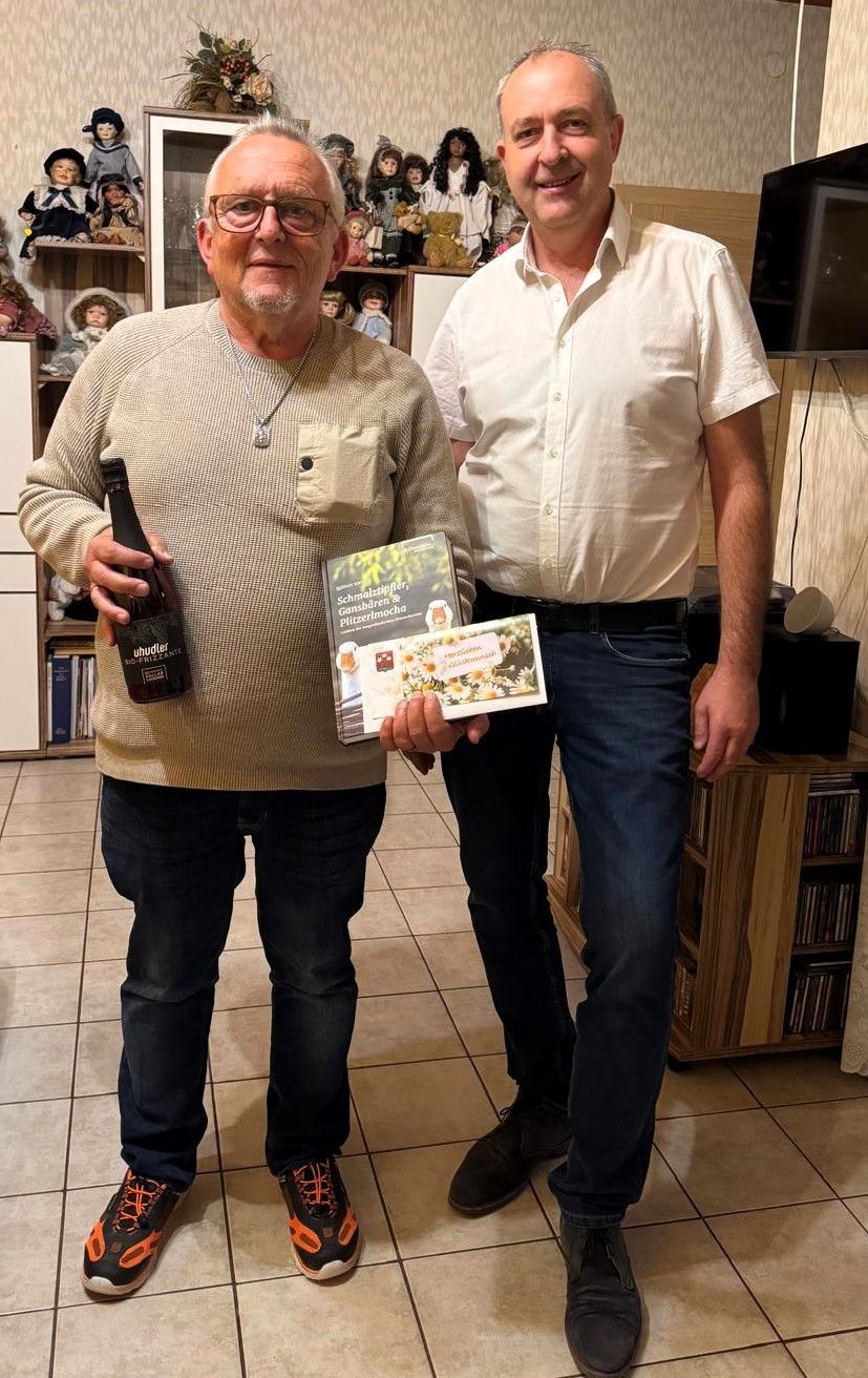 Zwei Männer stehen in einem Raum, einer hält ein Buch und eine Flasche, der andere ein Buch. Sie lächeln beide. Hinter ihnen sind Regale mit Puppen, ein Fernseher und ein Schrank mit CDs. Der Boden ist gefliest.