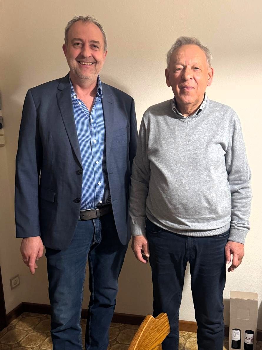 Zwei Männer stehen nah beieinander und lächeln gegen eine helle Wand. Der Mann links trägt einen blauen Blazer über einem karierten Hemd, und der Mann rechts trägt einen grauen Pullover.