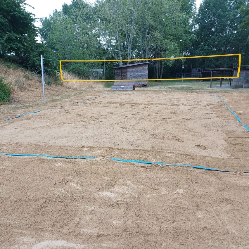 Ein Outdoor-Volleyballplatz mit einem gelben Netz und blauen Grenzlinien auf einem sandigen Untergrund, umgeben von Bäumen und einer Holzhütte.