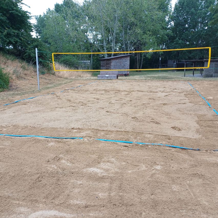 Ein Outdoor-Volleyballplatz mit einem gelben Netz und blauen Grenzlinien auf einem sandigen Untergrund, umgeben von Bäumen und einer Holzhütte.