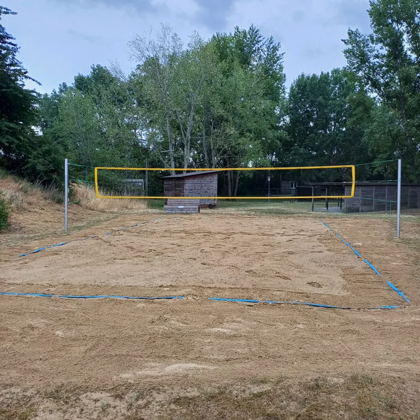 Ein Freiluft-Volleyballplatz mit einem gelben Netz und blauen Grenzlinien auf einem sandigen Feld, umgeben von Bäumen und einem Holzschuppen.
