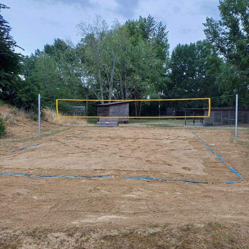 Ein Freiluft-Volleyballplatz mit einem gelben Netz und blauen Grenzlinien auf einem sandigen Feld, umgeben von Bäumen und einem Holzschuppen.