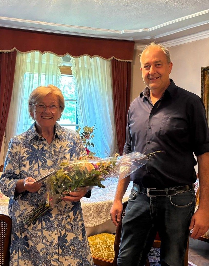 Ein Mann und eine Frau stehen in einem Raum mit einem Tisch und Blumen im Vordergrund. Die Frau hält einen Blumenstrauß und lächelt, während der Mann einen Blumenstrauß hält und sie ansieht. Hinter ihnen ist ein Fenster mit Vorhängen und ein gerahmtes Bild an der Wand.