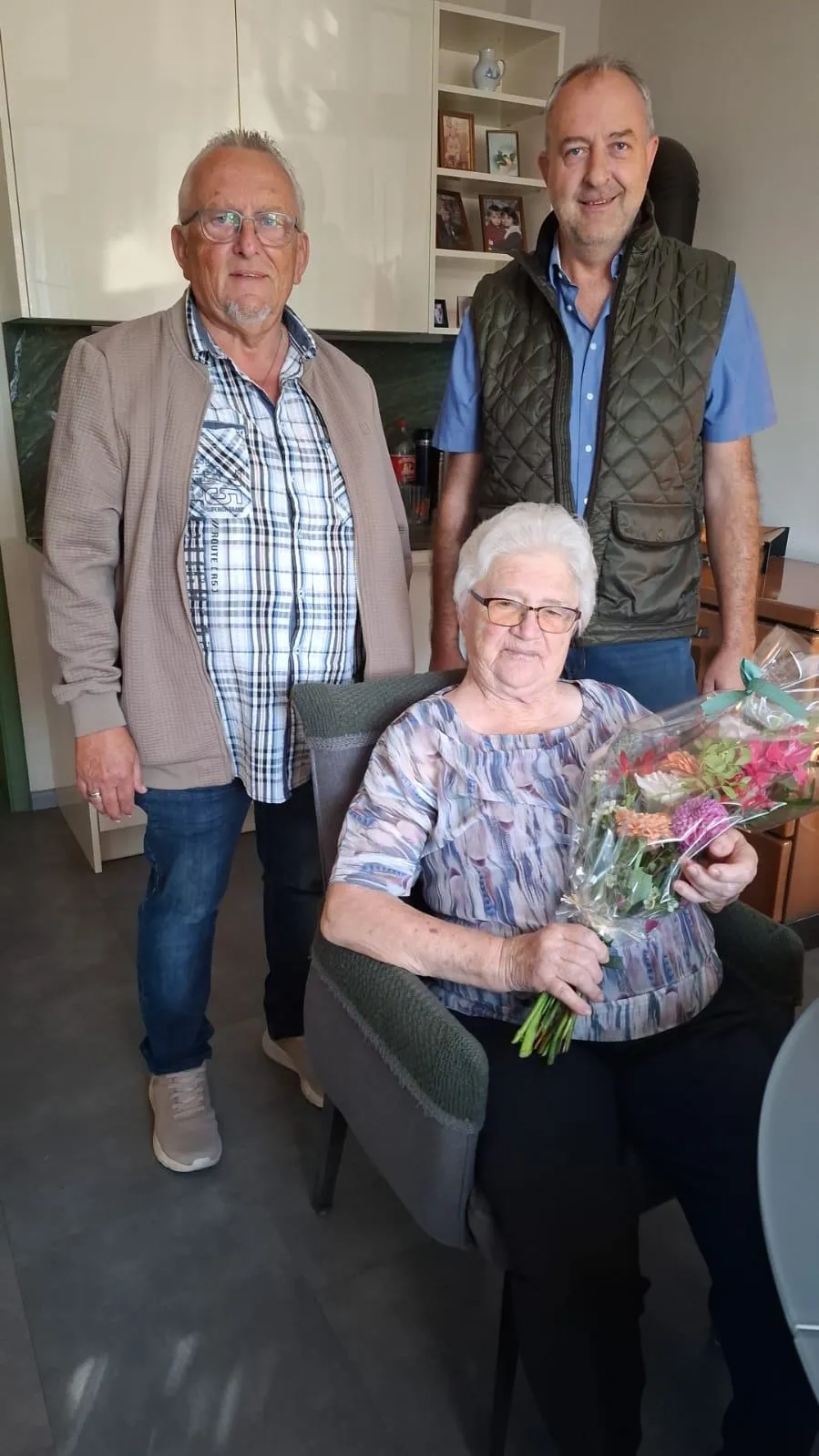 Eine ältere Frau mit weißen Haaren sitzt in einem Stuhl und hält einen Blumenstrauß. Zwei Männer stehen hinter ihr, einer trägt ein kariertes Hemd und der andere eine Weste.