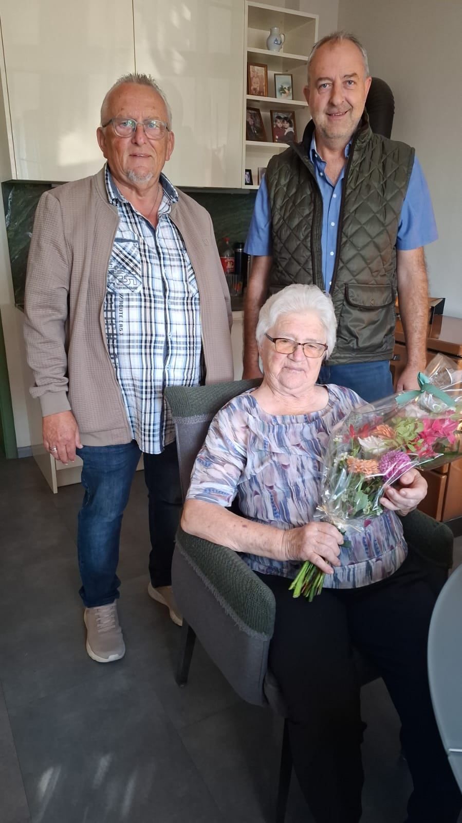 Eine ältere Frau mit weißen Haaren sitzt in einem Stuhl und hält einen Blumenstrauß. Zwei Männer stehen hinter ihr, einer trägt ein kariertes Hemd und der andere eine Weste.