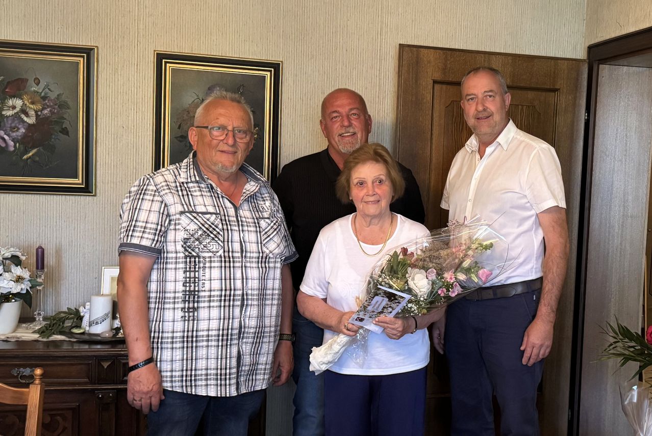 Vier Menschen stehen in einem Raum mit einer Blumenwanddekoration. Eine ältere Frau hält einen Blumenstrauß. Ein Mann in einem karierten Hemd lächelt. Ein anderer Mann trägt ein schwarzes Hemd.