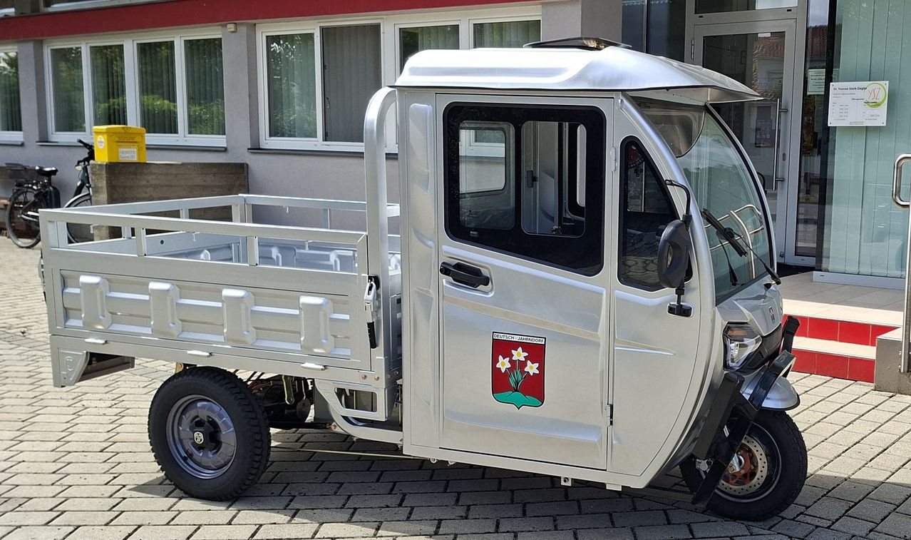 Ein silberner Cargo-Dreirad mit offener Tür und einem roten Logo mit Blumen vor einem Haus geparkt.