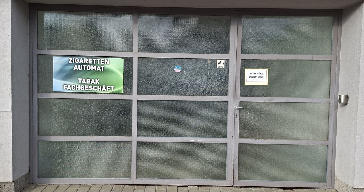 Eine Glastür mit mehreren Paneelen, einschließlich eines grünen Schilds mit 'RETTEN OMA', einem CCTV-Schild und einem weißen Schild mit 'BITTE TÜRE SCHLIESSEN!!!!'. Die Tür ist geschlossen.