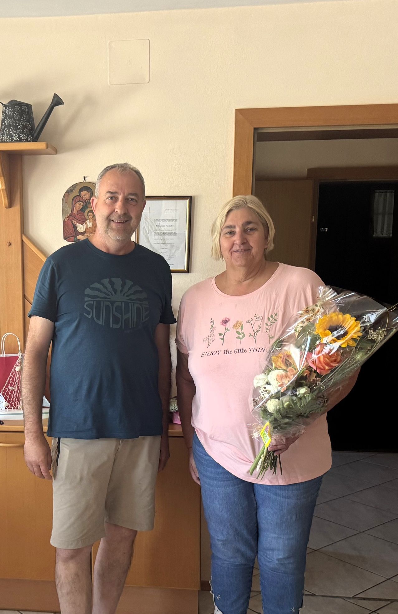 Ein Mann und eine Frau stehen eng beieinander und lächeln. Der Mann trägt ein blaues T-Shirt mit dem Wort 'SUNSHINE'. Die Frau hält einen Blumenstrauß. Hinter ihnen ist eine Holzwand mit einem gerahmten Bild und einem Regal.