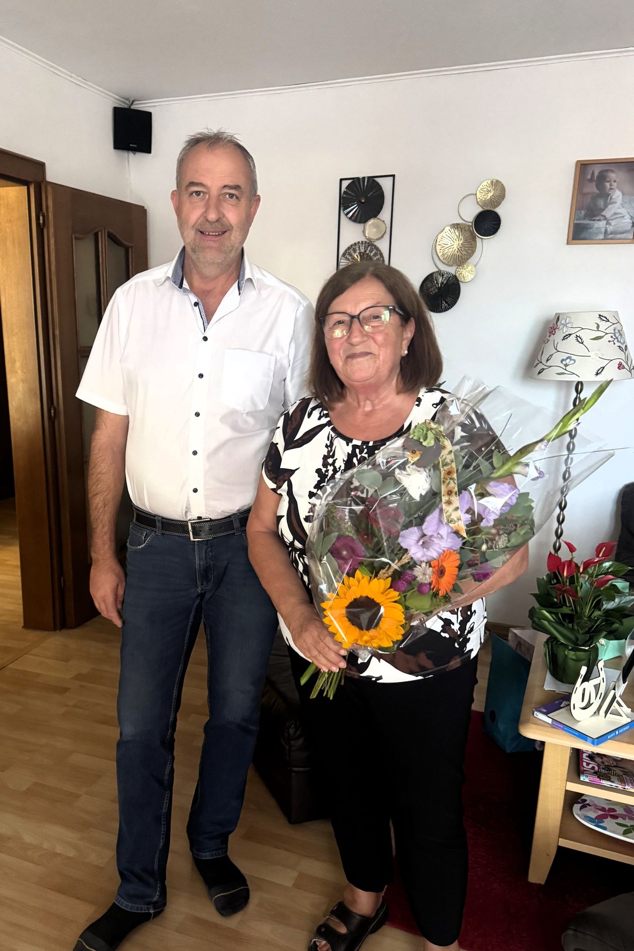 Ein Mann und eine Frau stehen gemeinsam in einem Raum, die Frau hält einen Blumenstrauß. Hinter ihnen ist eine weiße Wand mit dekorativen Gegenständen.